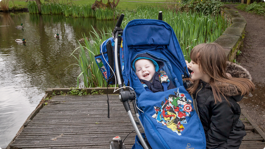 Child in blue buggy with older sibling, standing by pond