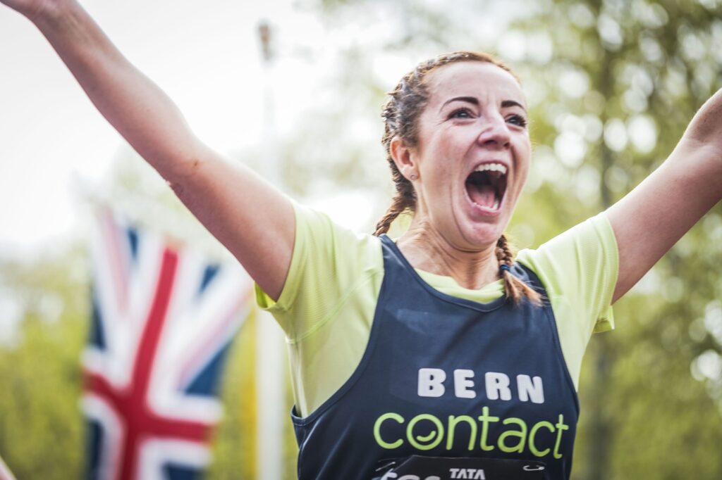 A woman with plaited hair and a race number smiles with her arms raised in celebration. A blurred Union Jack flag appears in the background, suggesting a race event.