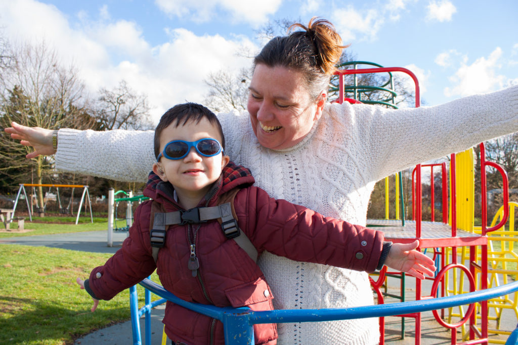 A woman and a young boy stand on playground equipment, both smiling and spreading their arms wide in a playful manner. The boy wears sunglasses and a red coat, while the woman sports a white sweater. The background includes a clear sky and additional playground structures.