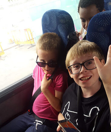 Two boys with short hair sit next to each other on a bus in seats with blue upholstery. Both are wearing glasses and seatbelts. One boy in a pink shirt is smiling and holding his glasses. The other boy in a black shirt is smiling and holding a smartphone.