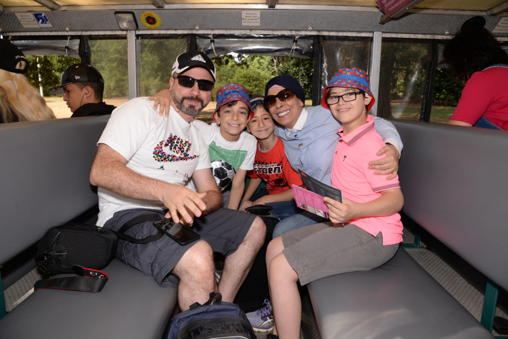A family of five sits closely together on a bus. The parents, both wearing sunglasses and caps, sit on either end and smile at the camera. The three children, dressed in colorful hats, look happy and hold electronic devices. Trees can be seen through the bus windows.