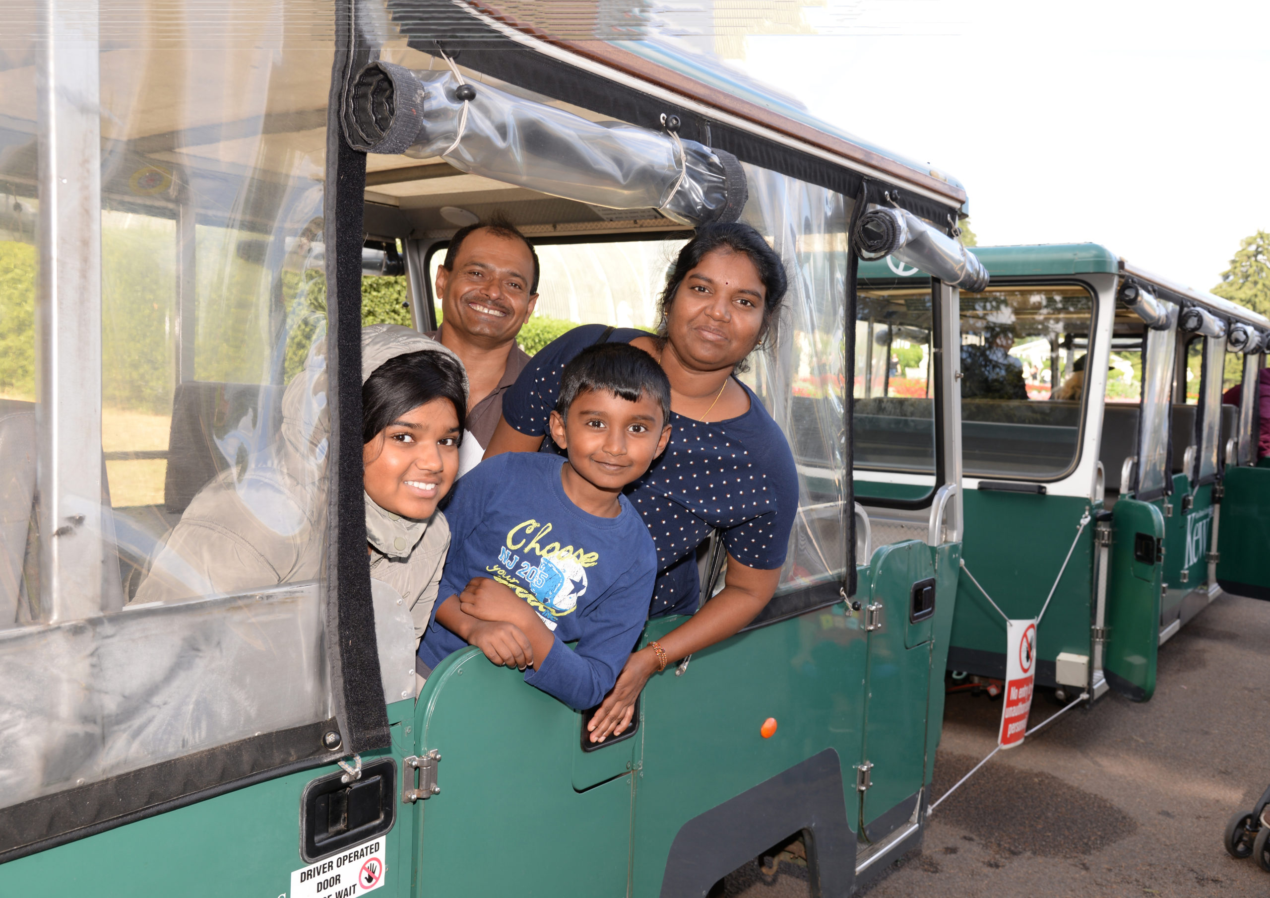 A family of four, including two adults and two children, leans out from the windows and doorway of a green tram or train-like vehicle. They are smiling and appear to be enjoying their time, with greenery visible in the background.