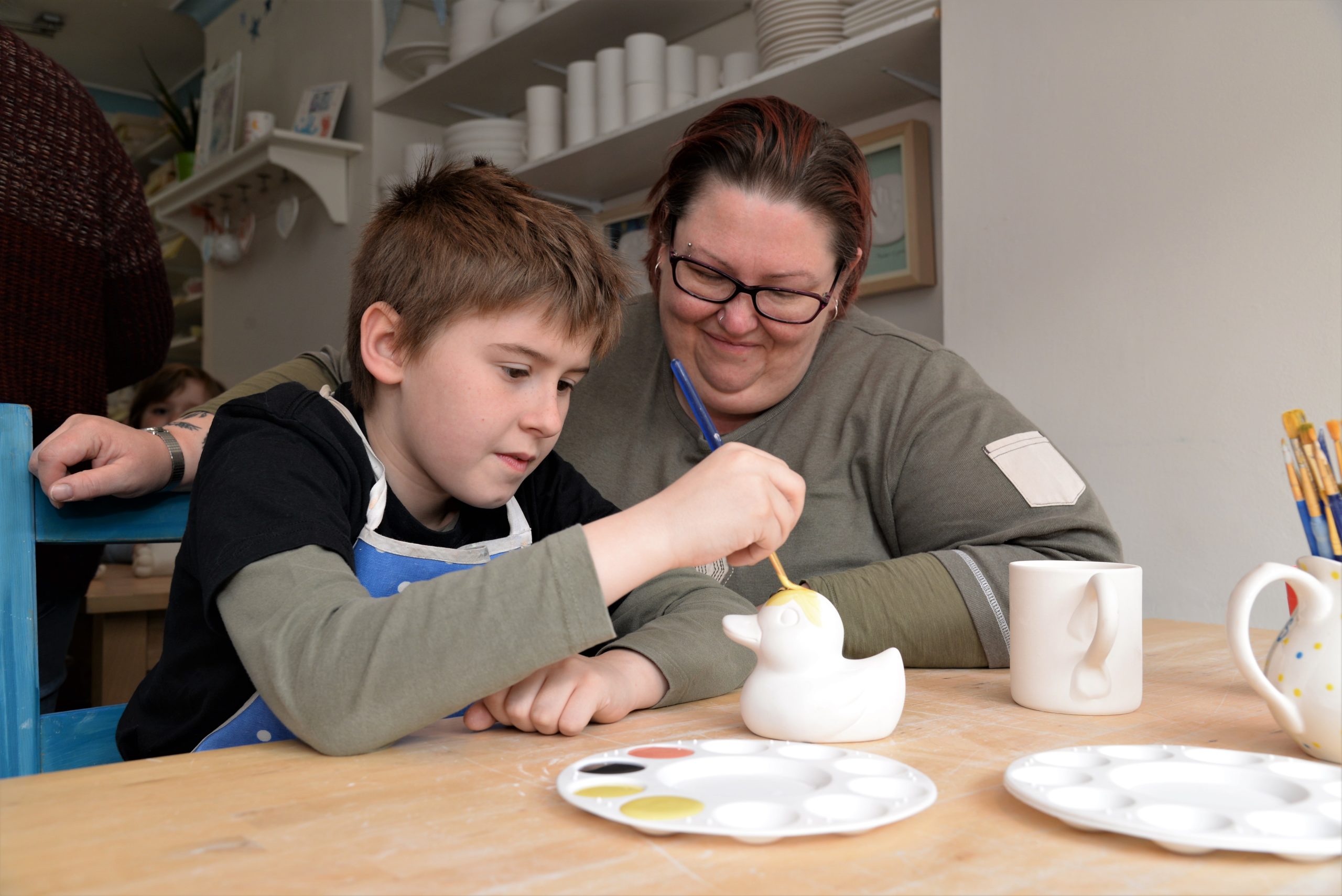 A child and an adult sit at a table in a pottery studio, painting a ceramic duck. The boy is focused on his work, holding a paintbrush and applying yellow paint to the duck while the adult smiles and watches. Paint palettes and other ceramic items are on the table.