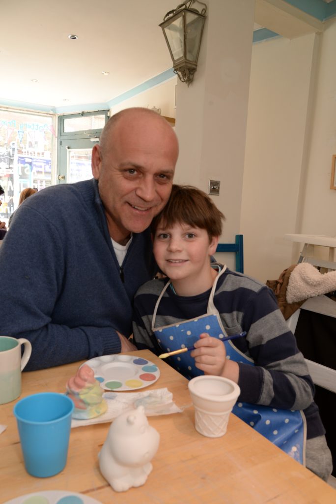 A smiling man and a boy are sitting at a table in a bright room, engaged in painting pottery. The man wears a blue sweater, and the boy wears a striped sweater and a blue apron. Various painting supplies and ceramics are on the table in front of them.