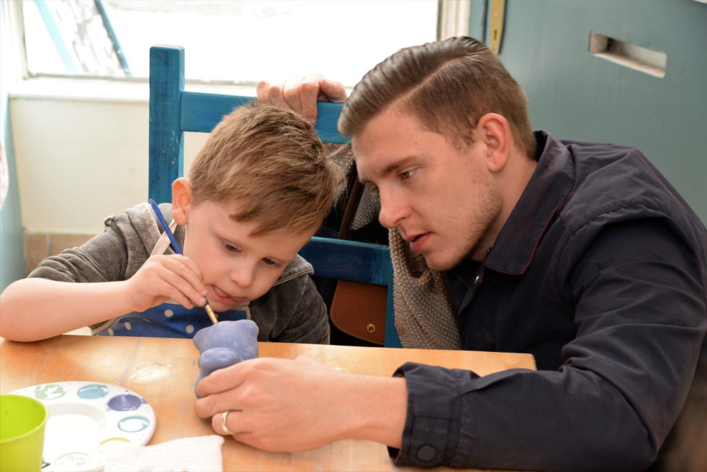 A young boy is carefully painting a small ceramic object, focusing intently with a paintbrush in his hand. A man, sitting beside him, is guiding and observing the boy's work, offering support. They are seated at a wooden table with paints and brushes scattered around.