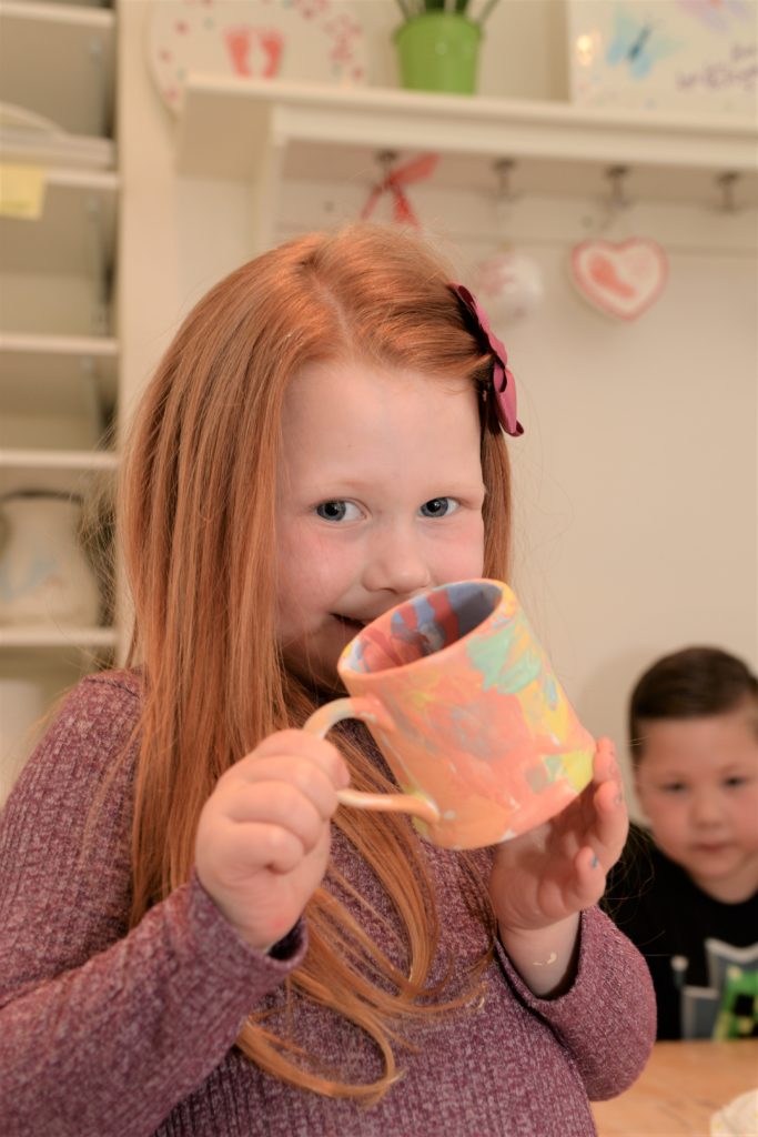 A young girl with long red hair and a bow smiles while holding a colorful painted mug in a pottery workshop. She wears a purple sweater. In the background, a boy and shelves with various pottery and decorations are visible.