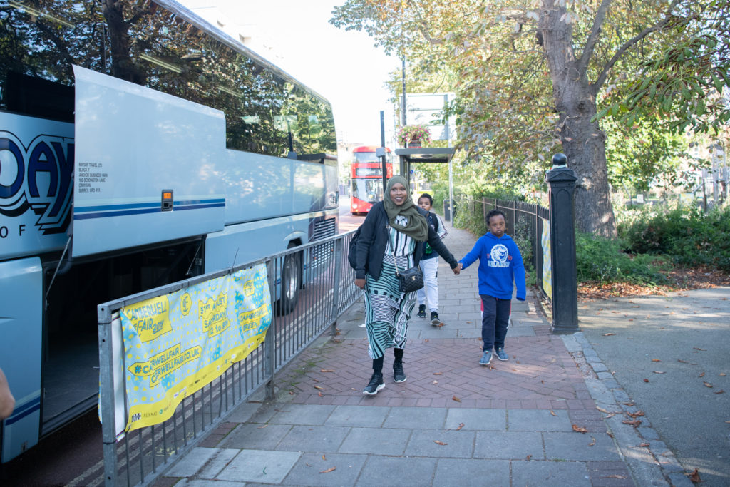 A woman wearing a hijab and a young boy in a blue hoodie hold hands as they walk along a sidewalk. A large white coach bus is parked on the street. Trees with green and autumn-colored leaves line the background. A yellow banner is attached to a nearby railing.