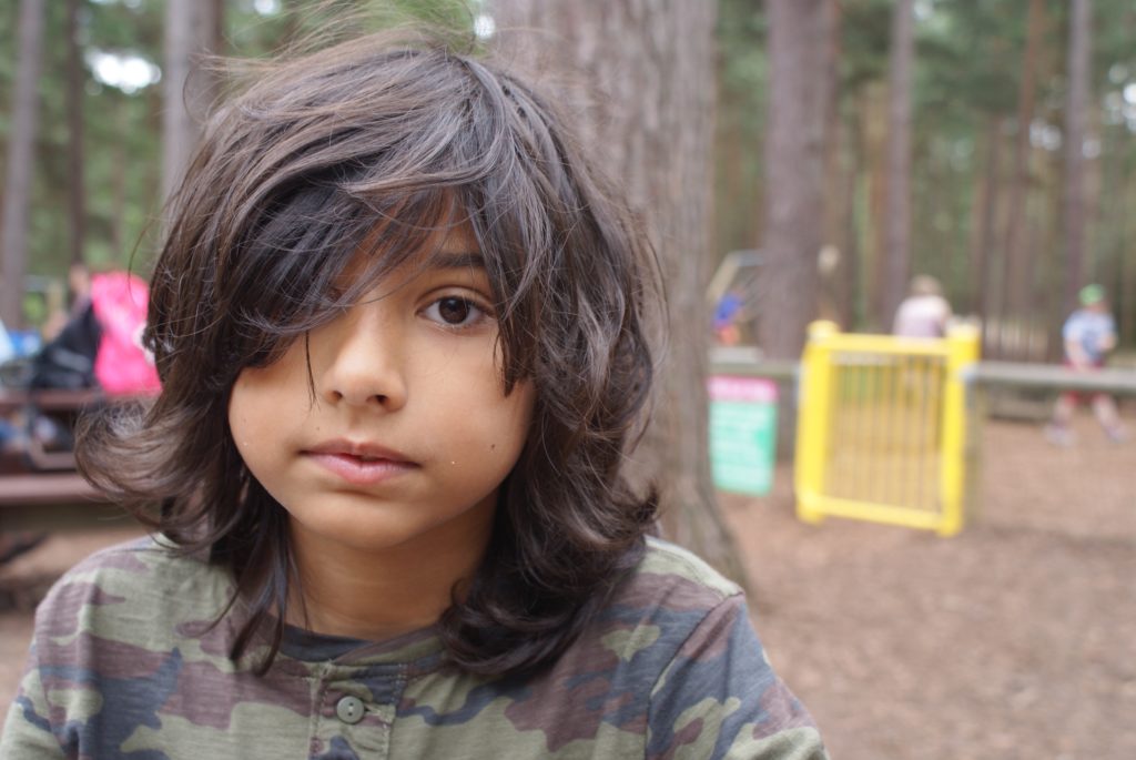 A young child with long, dark hair is wearing a camouflage shirt and is looking directly into the camera. In the background, there are blurred trees and people, suggesting an outdoor park or forest setting. A yellow gate is also visible.