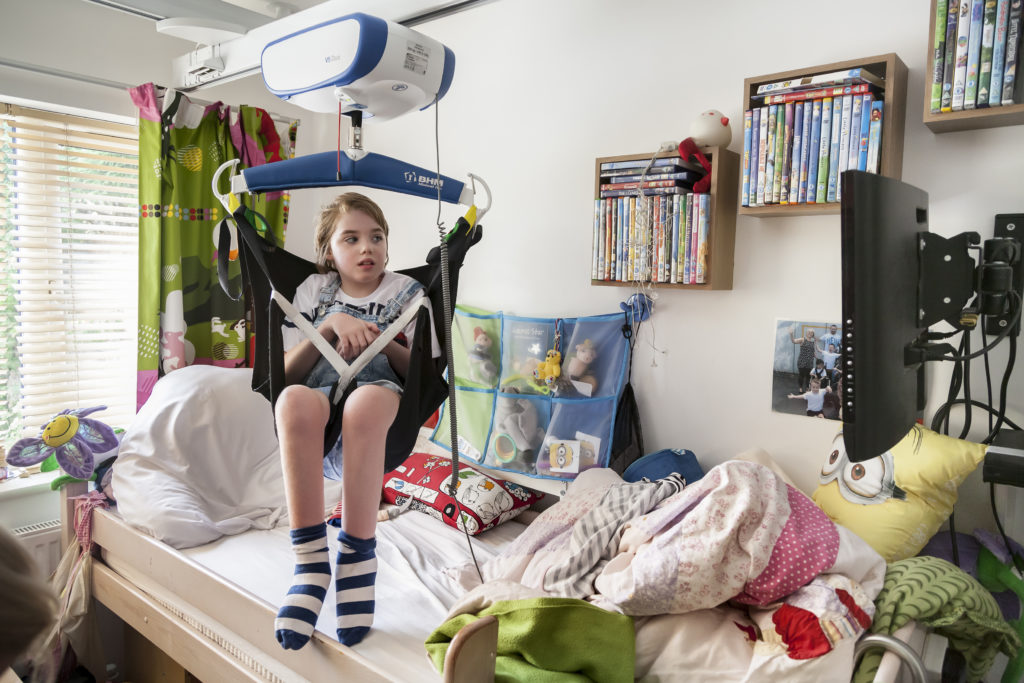 A young child with striped socks sits in a sling attached to a ceiling lift above their bed in a cozy, colorful room. The room is decorated with stuffed animals, DVDs on shelves, and vibrant bedding. The child appears focused and engaged.