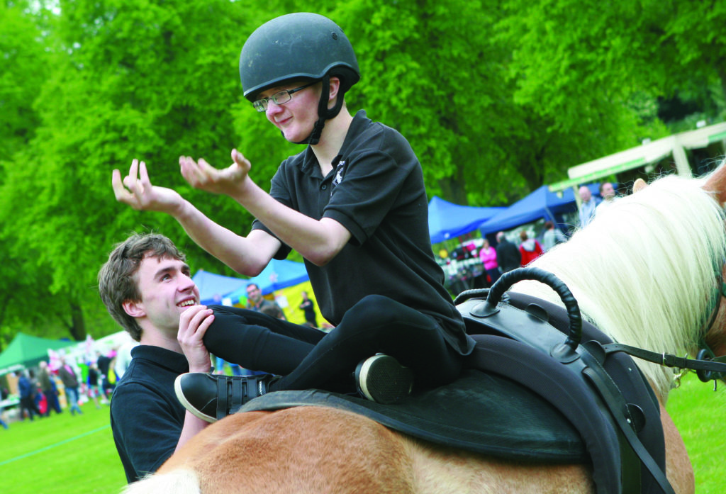 A smiling person wearing a helmet sits on a horse, holding their hands up, assisted by another smiling person who is holding their leg. Both are dressed in black shirts. The background features a green park with trees and several tents or booths.