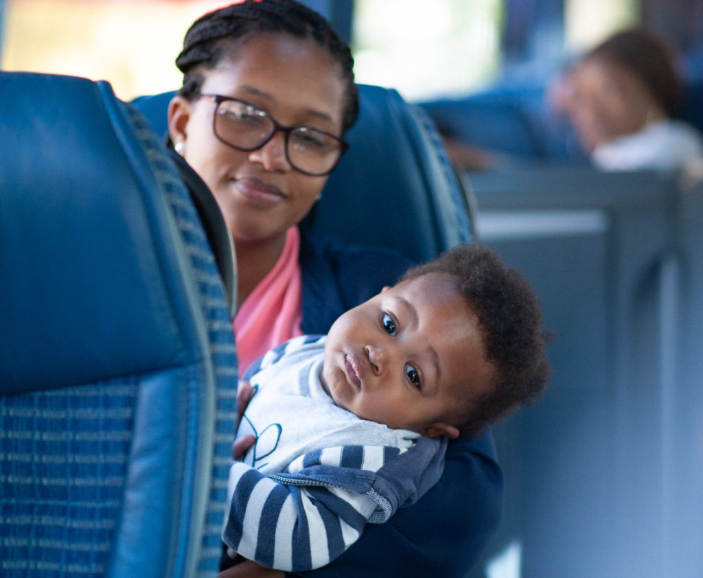 A woman with glasses and braided hair holds a baby dressed in a striped shirt while sitting on a blue fabric seat in a vehicle, likely a bus or train. The baby looks curiously at the camera. The interior is softly lit, and other passengers are blurred in the background.