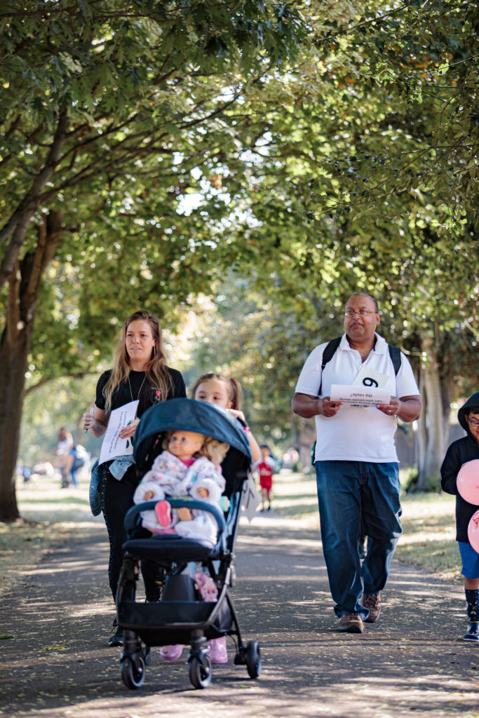 A group of people walks down a tree-lined path on a sunny day. A woman is pushing a stroller with a child, while a man holds papers. Another child is holding a balloon, and various other people are visible in the background. The atmosphere is calm and leisurely.