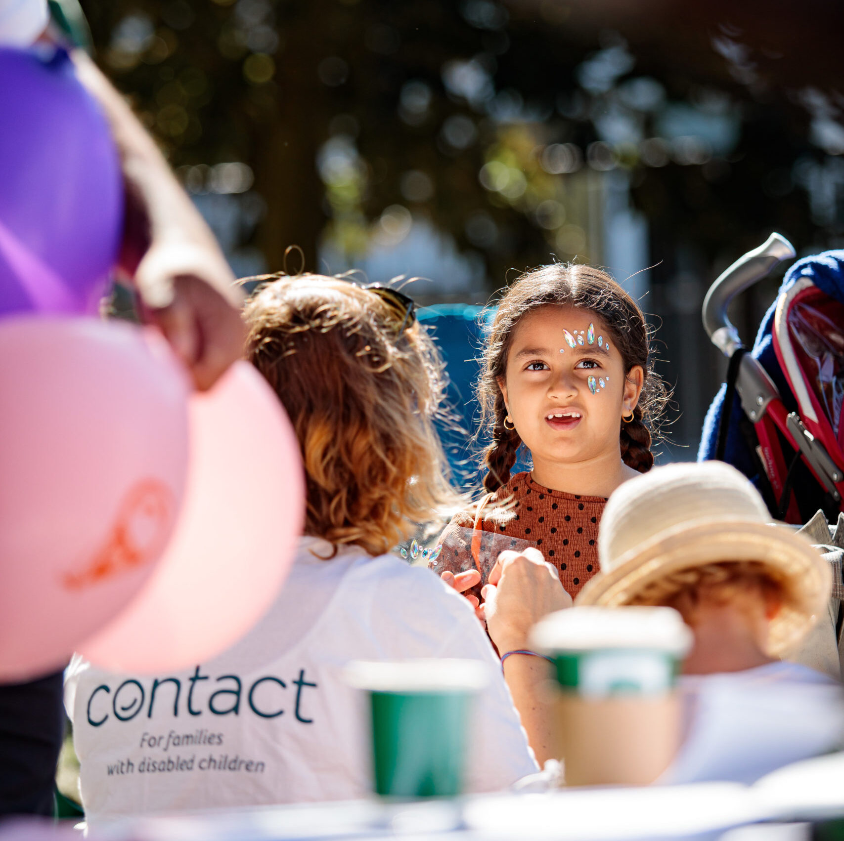 A child with face paint talks to an adult at an outdoor event. The adult wears a white "Contact" t-shirt for a charity supporting families with disabled children. There are balloons, cups, and blurred people in the background, indicating a lively atmosphere.