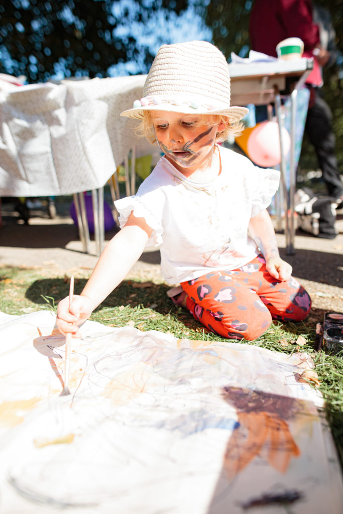 A young child wearing a straw hat and white shirt is kneeling on the grass, painting on a large piece of paper on the ground. The child has paint on their face and clothes. Tables with art supplies and balloons are visible in the background.