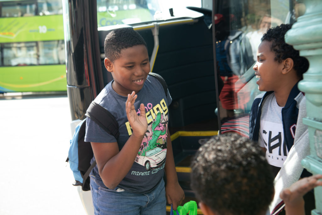 A young boy smiles and waves as he steps off a bus with a backpack. Another boy stands nearby, looking at him, while a third child is seen from behind in the foreground. A green bus is visible in the background.