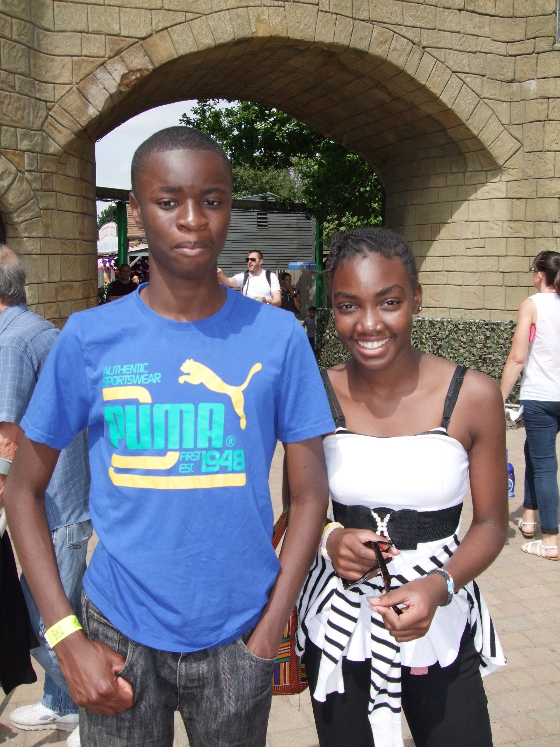 Two young people stand outdoors in front of a stone archway. The person on the left is wearing a blue PUMA t-shirt, and the person on the right is wearing a white and black striped top with a black belt. Both are smiling, and other people are visible in the background.