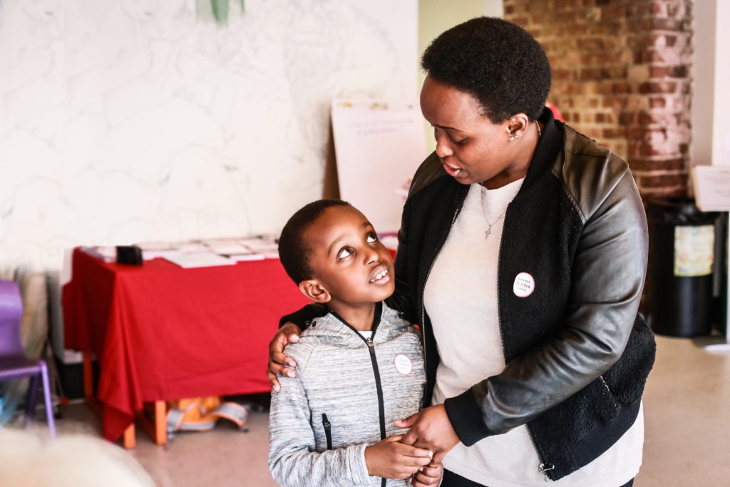 A woman with short hair and a black jacket embraces a young boy in a gray hoodie. They are indoors, standing in front of a red table with papers and a whiteboard. The boy looks up at the woman, and they both wear pin badges. The background includes a mural and brick column.