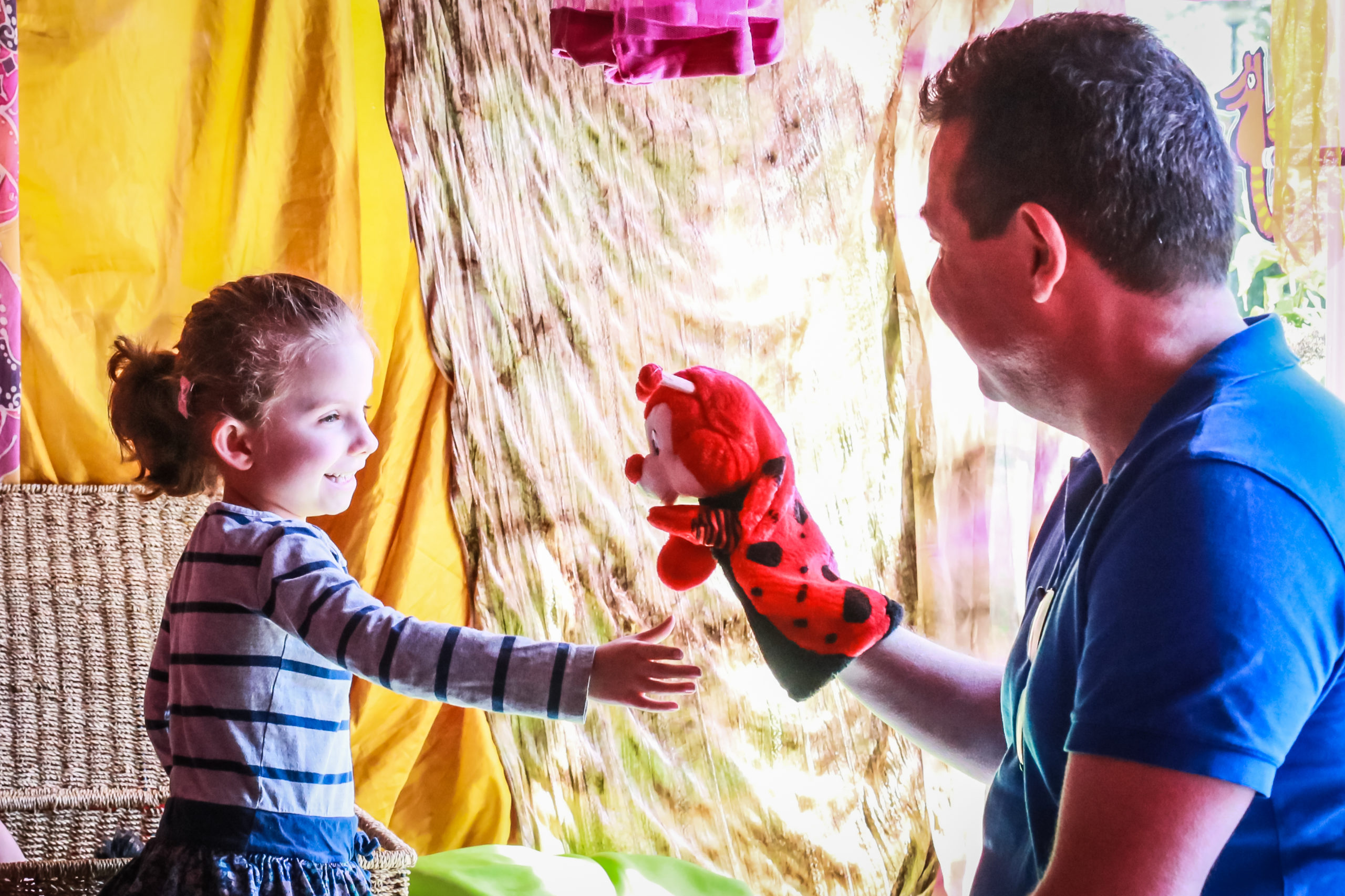 A young child with tied-back hair reaches out to touch a red hand puppet held by a smiling adult wearing a blue shirt. They are both sitting near colorful and patterned curtains, and the atmosphere appears joyful and playful.