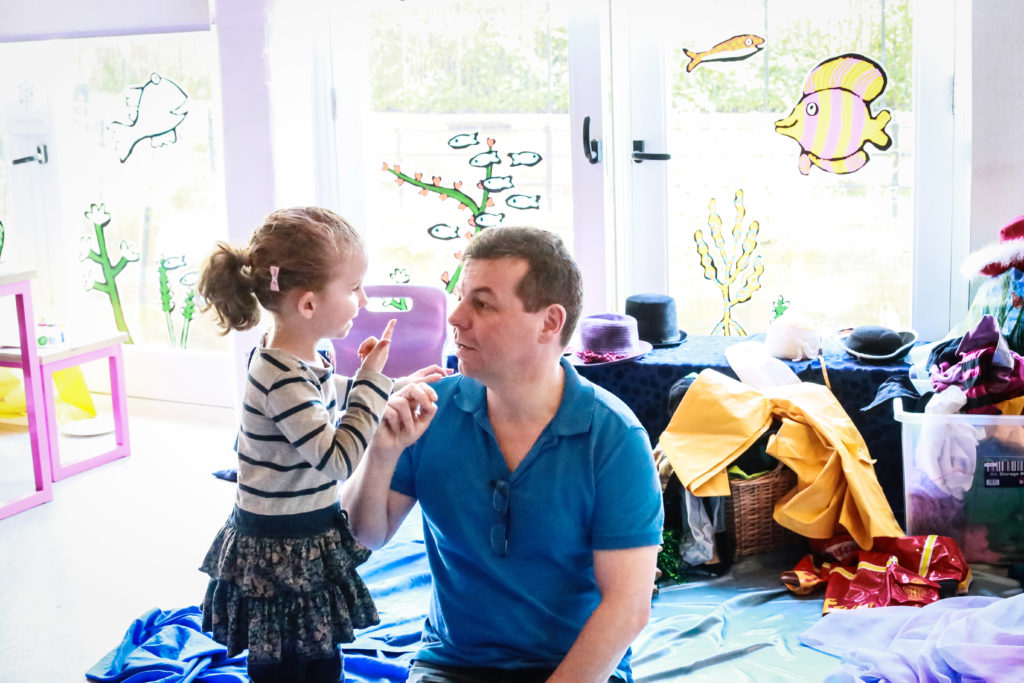 A man with short hair kneels on the floor, facing a young girl with curly hair who is standing. They are engaged in conversation. The room is decorated with colorful sea-themed stickers on the windows and scattered costumes and props around them.
