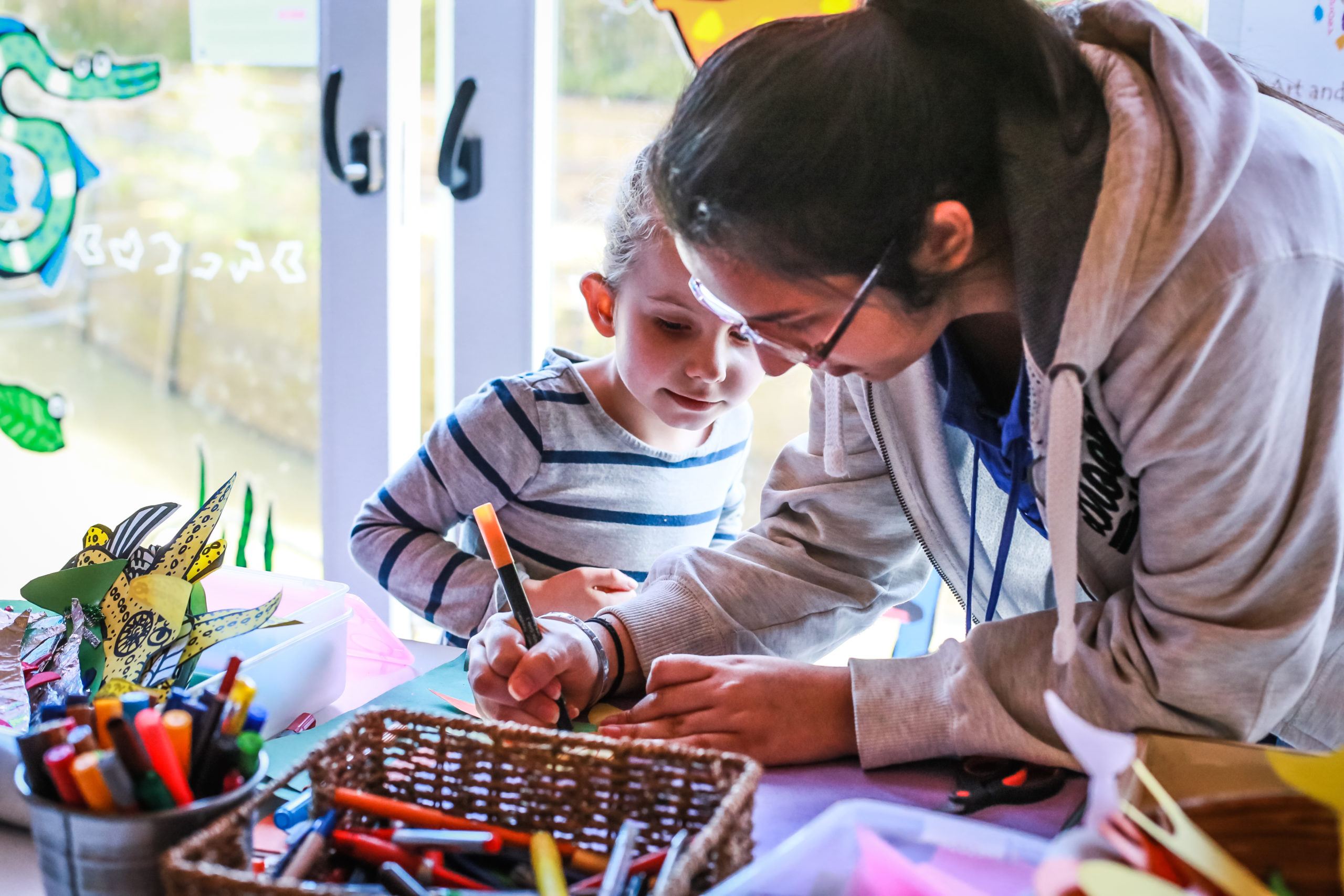 A young child and an adult are engaged in a craft activity at a bright indoor space. The child, in a striped shirt, is observing intently as the adult, in a hoodie, writes or draws on a piece of paper. Various colorful craft supplies are spread out on the table.