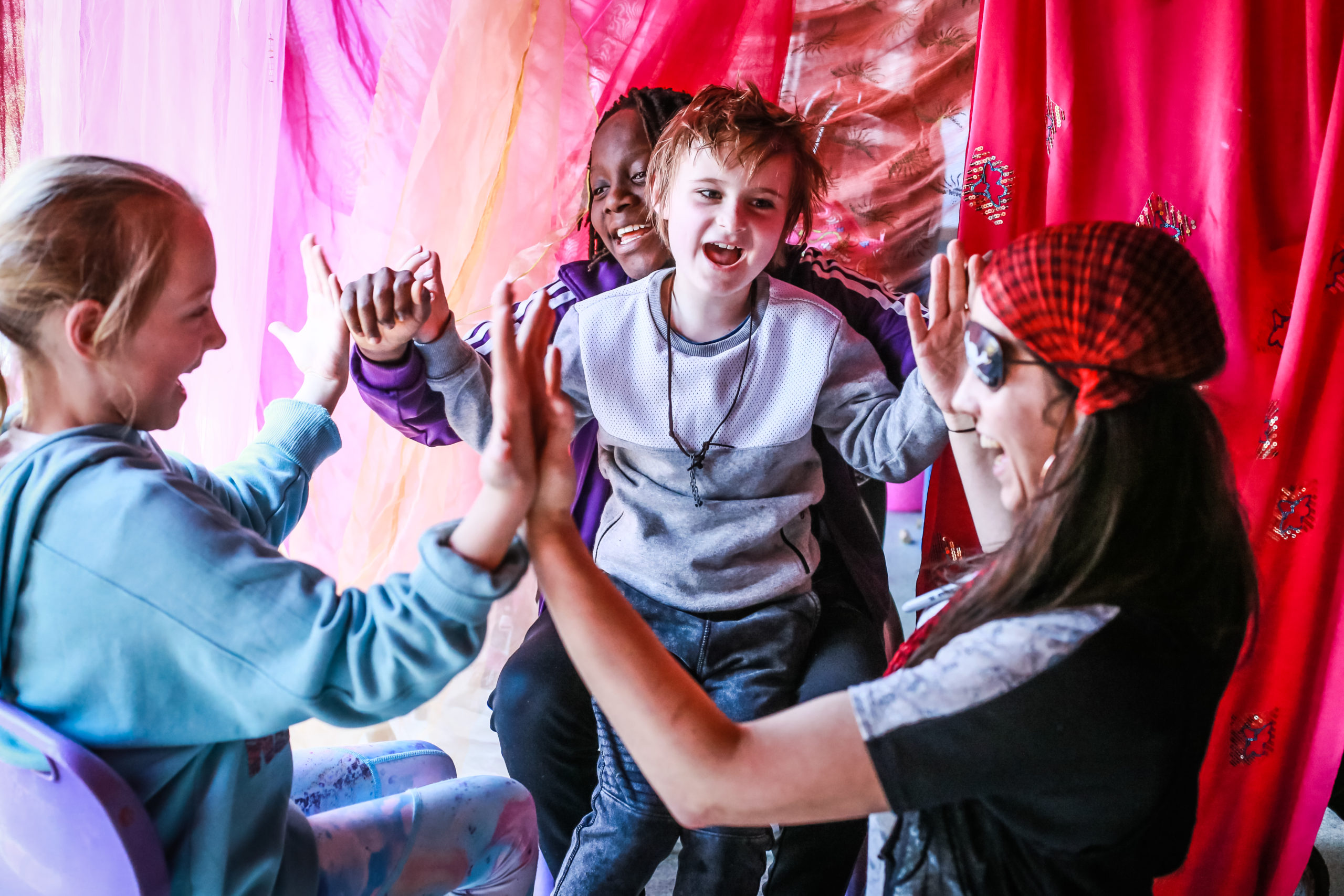 Four children are sitting in a colourful room draped with pink and red fabric, playing a clapping game. They appear joyful and engaged, with one child in the center smiling widely while others clap hands with each other, forming a circle.