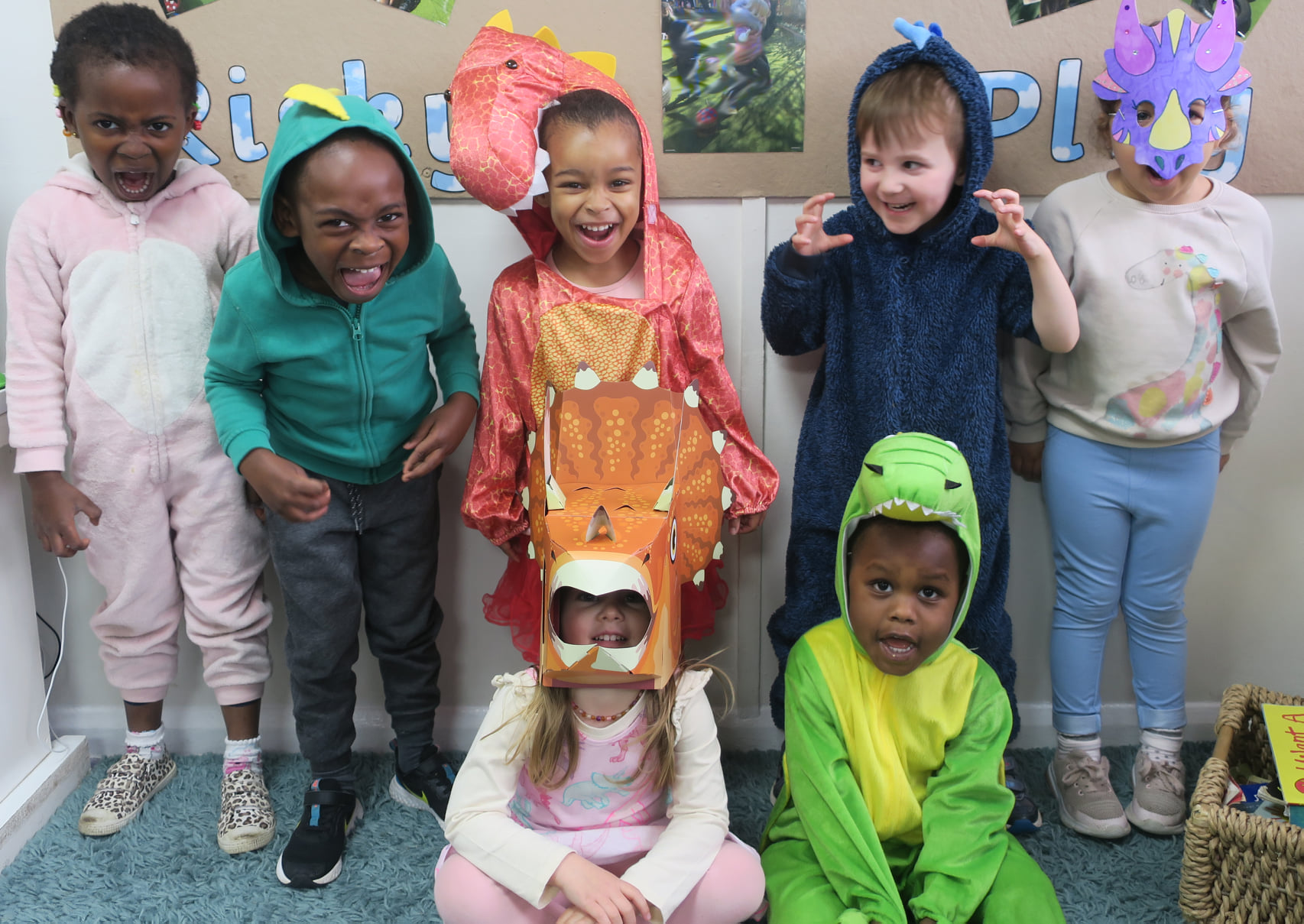 A group of seven young children dressed in various dinosaur costumes stand and sit together smiling and making playful, fierce poses. The background includes colorful artwork and decorations on a bulletin board. They appear to be in a classroom or play area.