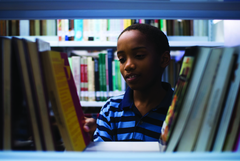 A young boy wearing a blue striped shirt browses for books on a shelf in a library. The bookshelves are filled with a variety of books, and the boy is focused on selecting a book. The scene is well-lit and colorful.