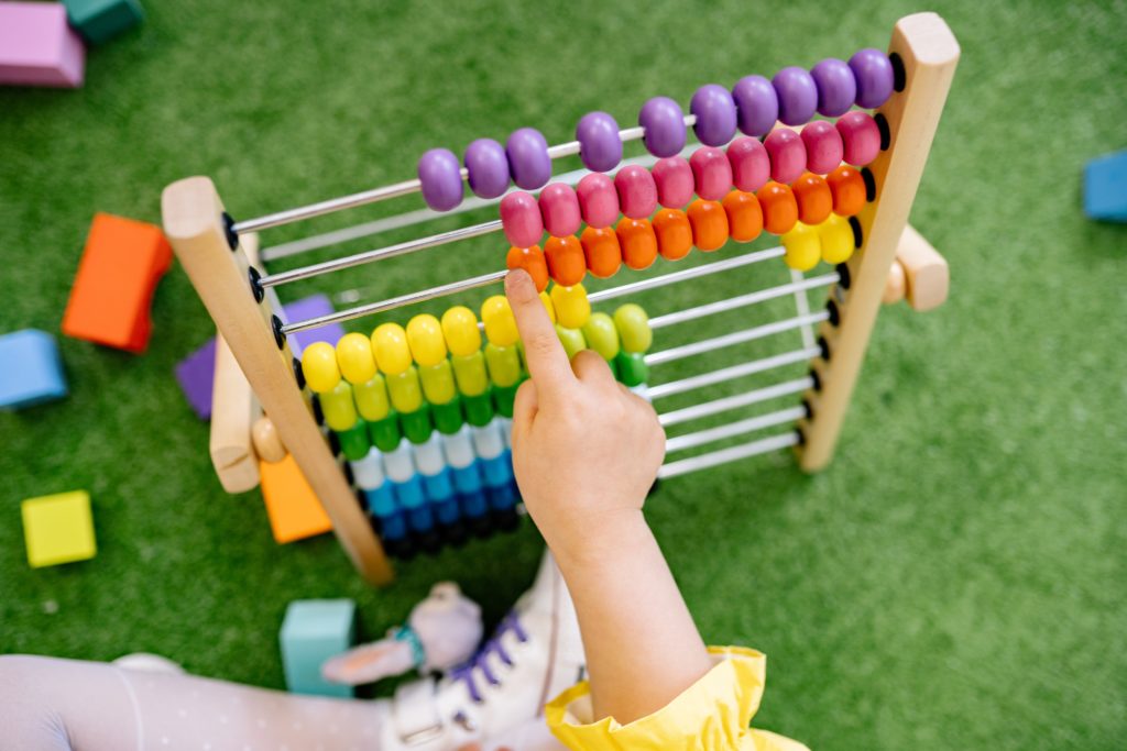 A child wearing a yellow sleeve points to the beads on a colorful abacus, which features rows of beads in red, orange, yellow, green, blue, and purple. Seated on a green carpet surrounded by scattered colorful blocks, the scene captures an engaging moment in early education.