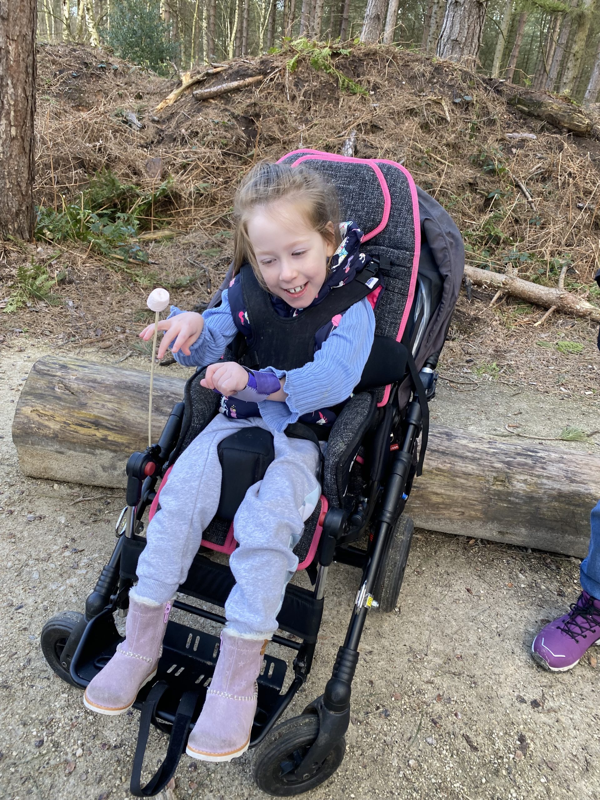 A smiling child sits in a black and pink wheelchair on a dirt path in a wooded area. She is holding a stick with a pink ball on top and appears to be in an outdoor, natural environment. A person's leg wearing jeans and trainers is partially visible on the right.