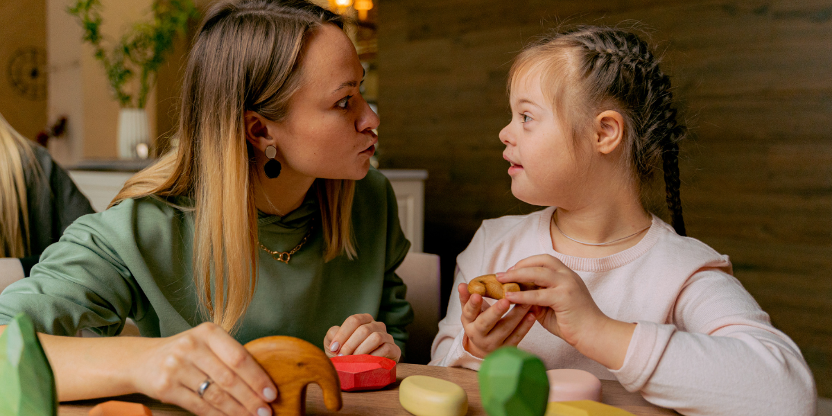 A young woman and a girl with Down syndrome are sitting at a table together. The girl is holding wooden toy blocks, and they are making eye contact. The woman appears to be speaking about school strikes while the girl listens intently in the cozy, indoor setting.