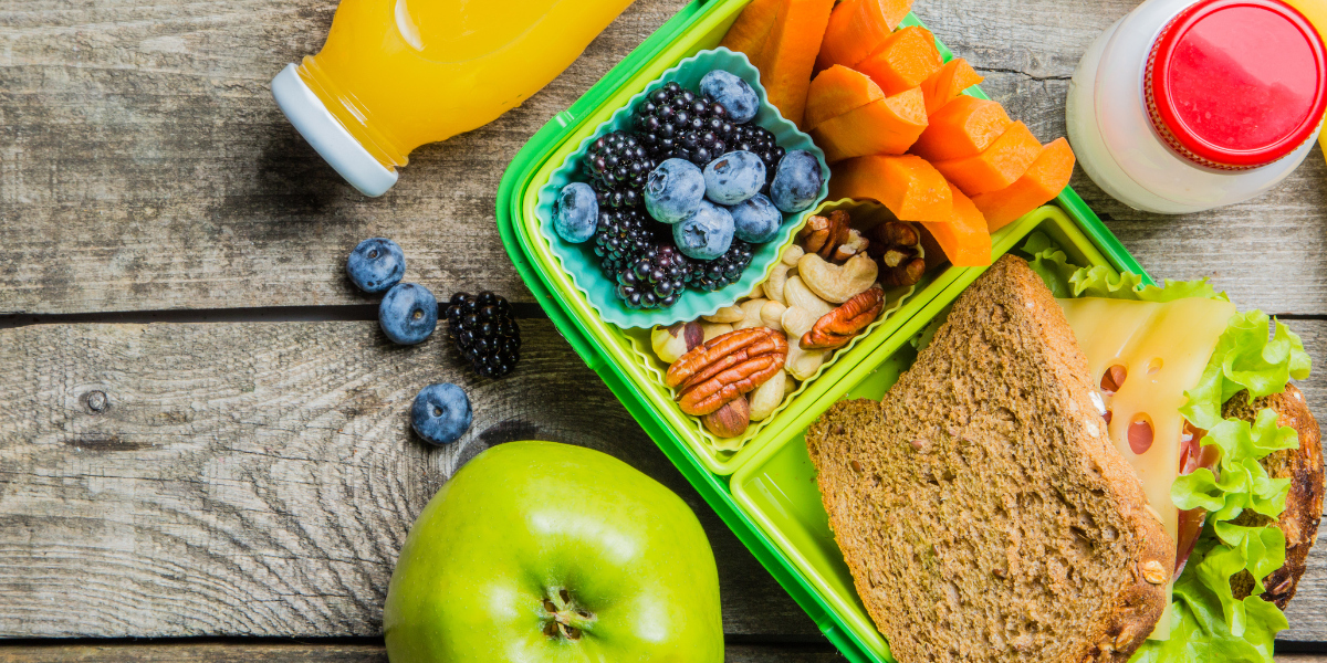 Healthy lunchbox on a wooden table filled with cheese sandwich, fruit, nuts, milk, orange juice and a green apple.