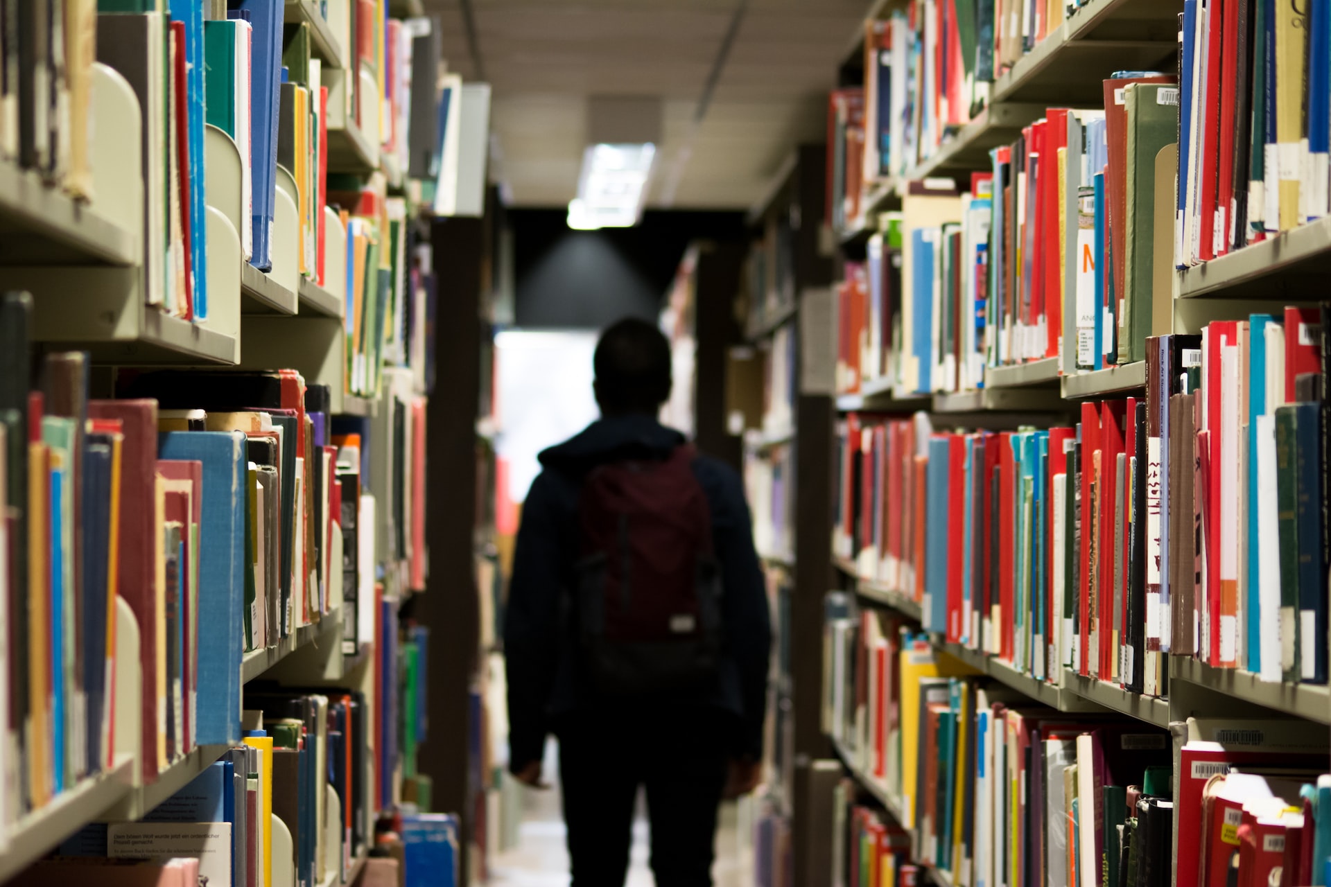 Student wearing a backpack, facing his back to the camera while standing between two bookshelves