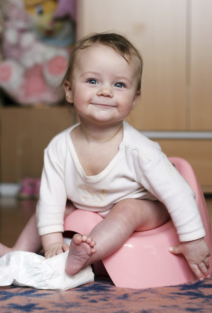 A smiling baby with light hair sits on a pink potty. Dressed in a white long-sleeved onesie, the baby holds a piece of white fabric or diaper. The background features a wooden cabinet and colorful toys, evoking a sense of growth similar to embarking on professional development courses. The baby appears content and playful.