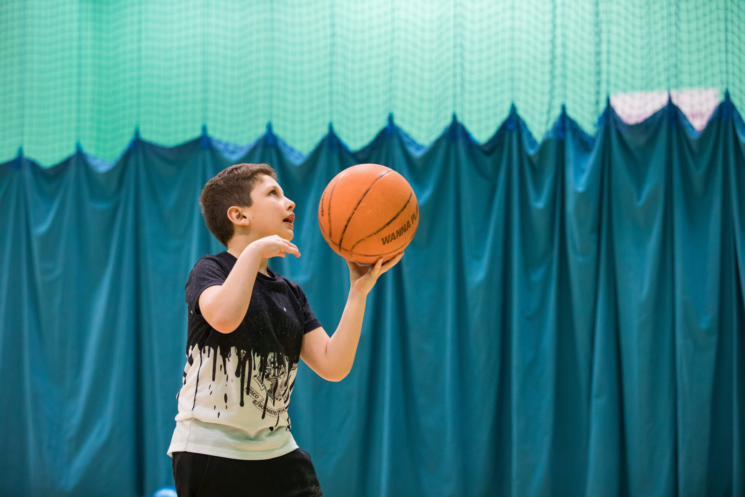 A young boy wearing a black and white shirt prepares to take a basketball shot in an indoor court, concentrating intently. A green curtain hangs in the background.