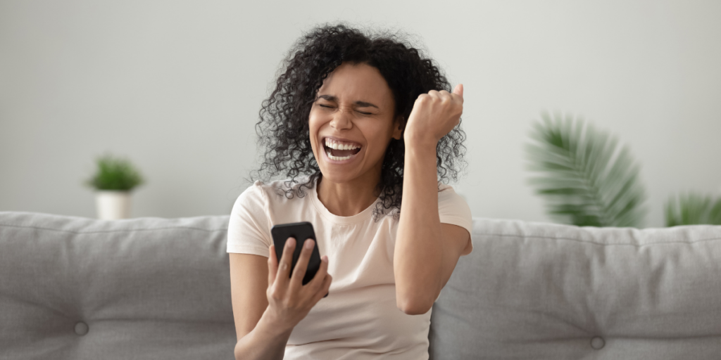 Women sitting on a sofa, celebrating after looking at her winning lottery numbers on her phone.