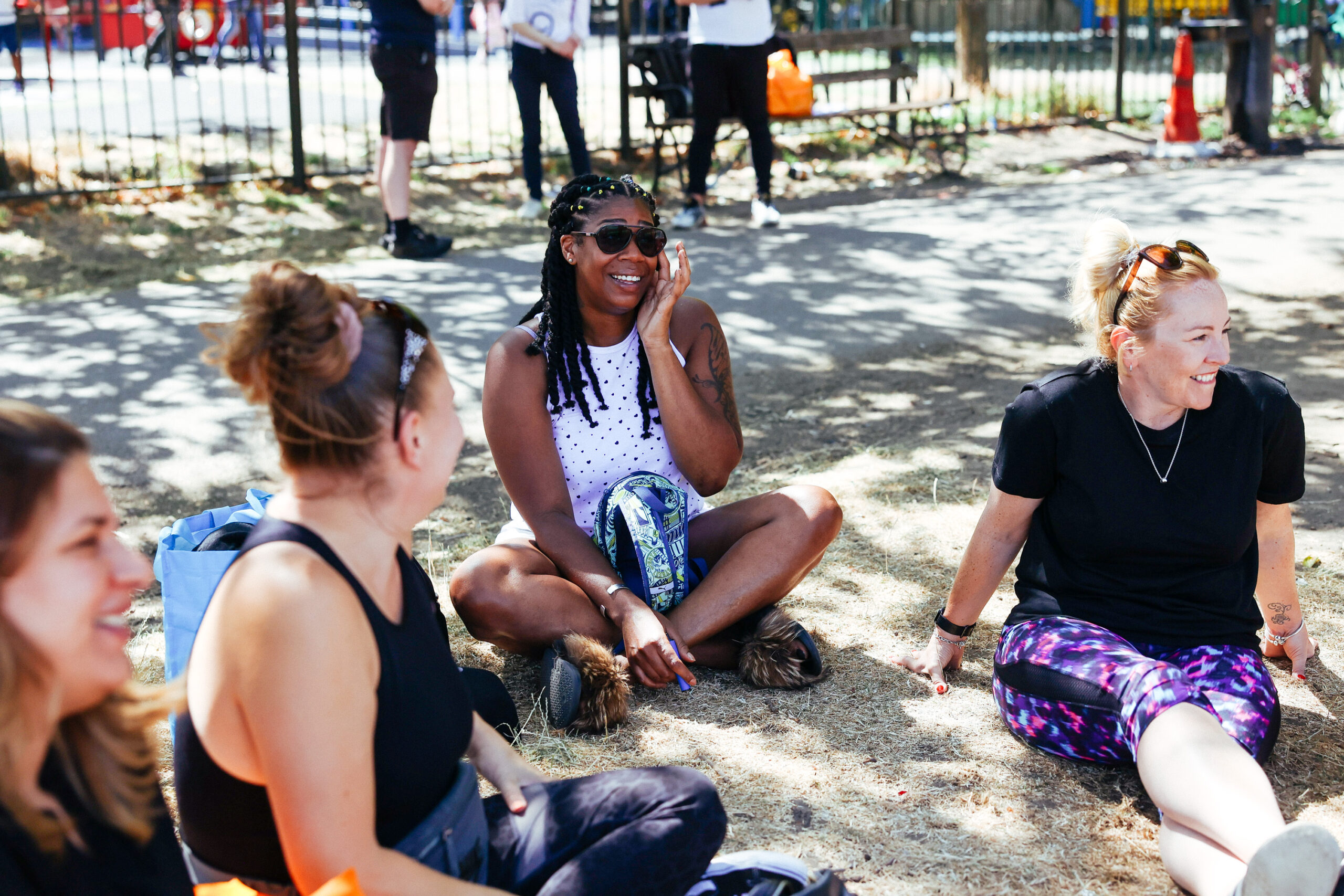 A group of friends sits on the ground in a park, laughing and enjoying a sunny day. They are casually dressed in athletic wear, and some are holding water bottles. Trees and a fence are visible in the background.