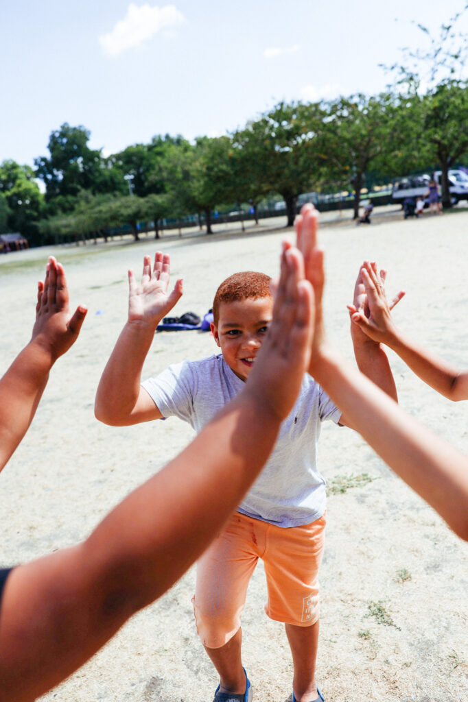 A young boy wearing a grey t-shirt and orange shorts is giving high fives to three other children with outstretched arms. They are outdoors in a park setting with trees in the background. The scene is bright and sunny.