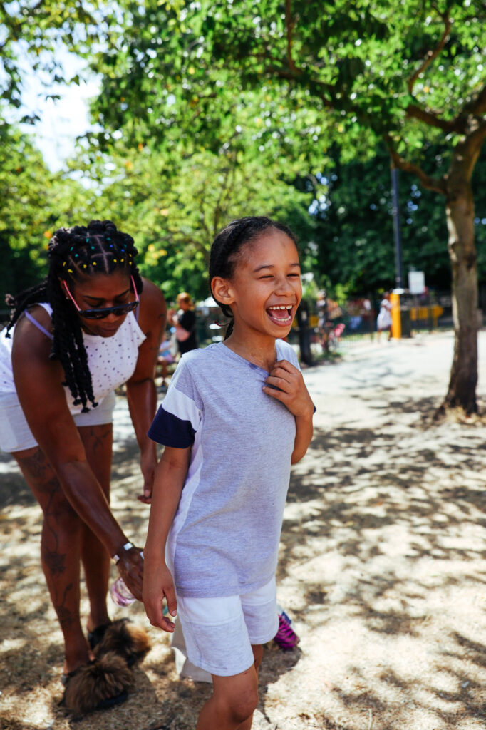 A woman in a white tank top and shorts playfully chases a laughing young child in a light gray shirt and white shorts through a sunny park. The scene is filled with trees and scattered sunlight, capturing a joyful moment.
