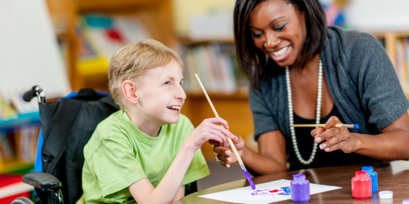 Teacher helping disabled child paint in a classroom setting - both are smiling.