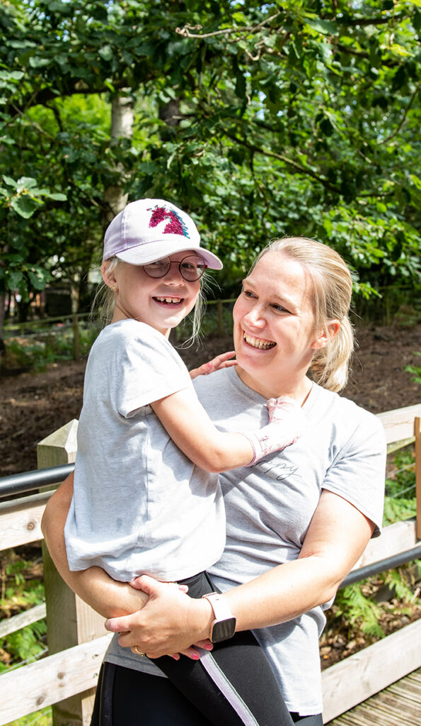 A smiling woman holding a young girl in her arms. The girl is wearing glasses, a pink cap, and a gray shirt, while the woman is dressed in a light gray shirt and has a smartwatch on her wrist. They are outdoors with greenery in the background on a sunny day.