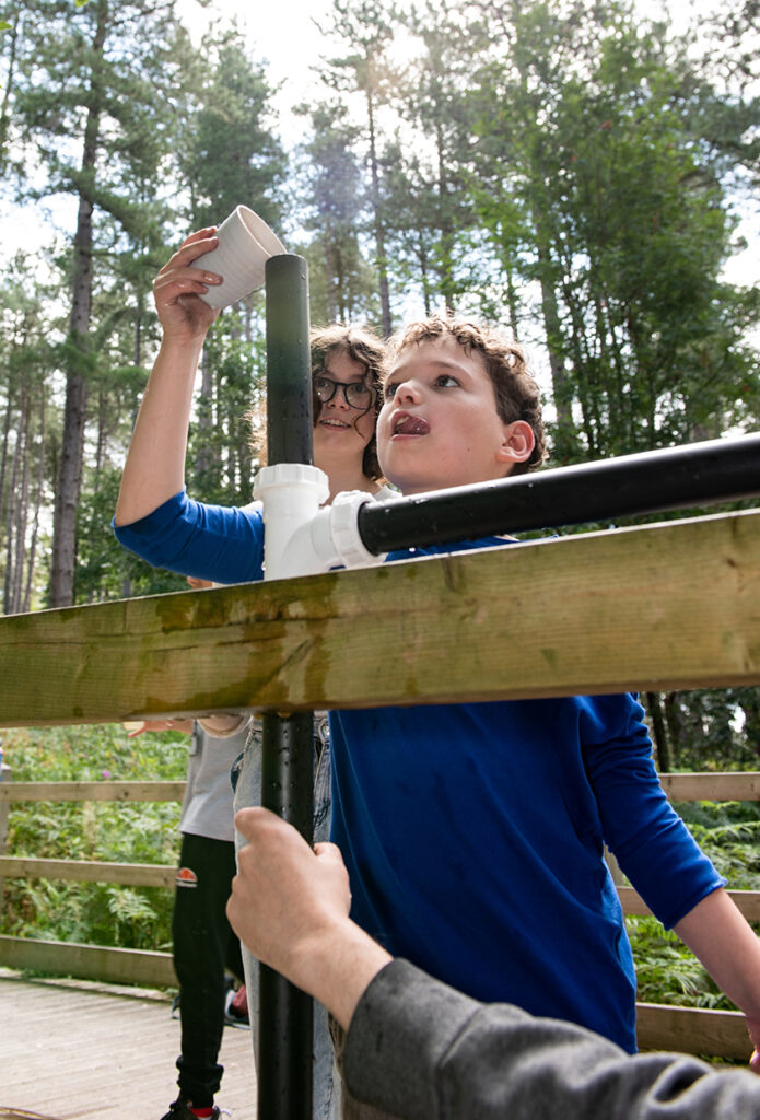 Two children are engaged in outdoor water play, interacting with a homemade PVC pipe water structure. One child holds a container above, while the other looks up at it intently. They are surrounded by a lush, forested area on a sunny day.