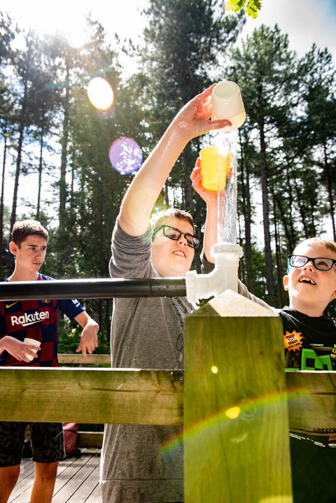 Three boys are working on an outdoor project with PVC pipes in a sunlit forest setting. One boy is wearing a gray shirt and glasses, reaching up to pour a yellow liquid into the pipe. Another boy, also with glasses, looks on. The third boy stands behind, observing.