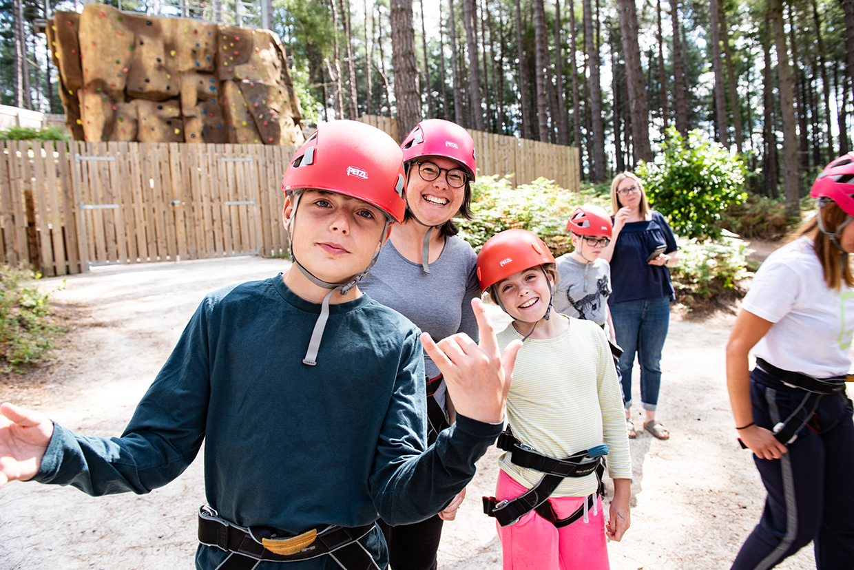 A group of people, including children and adults, wearing red helmets and climbing harnesses, are standing in an outdoor area with a wooden fence and a climbing wall in the background. They look happy, with one child making a hand gesture towards the camera.