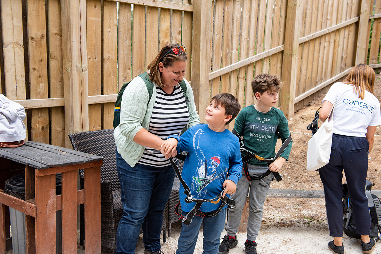 A woman helps a young boy fasten his safety harness, while another boy stands nearby, already wearing his harness. A person in a "centack" t-shirt is seen in the background. They are outdoors, by a wooden fence. Everyone appears to be preparing for an activity.