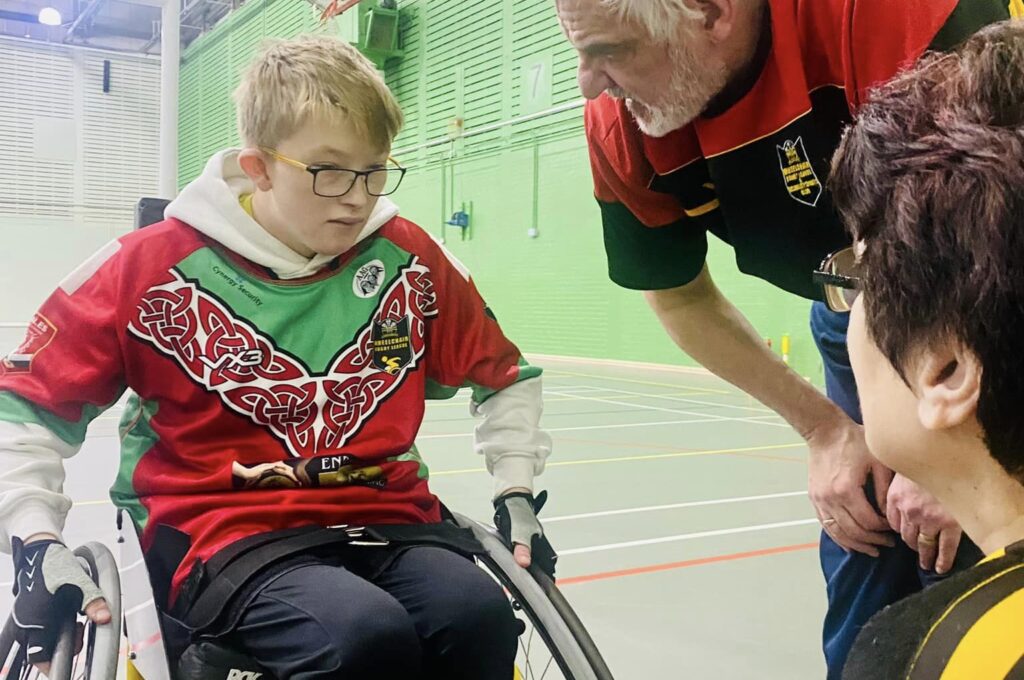Child playing wheelchair sports talkings to two adults in a gymnasium.