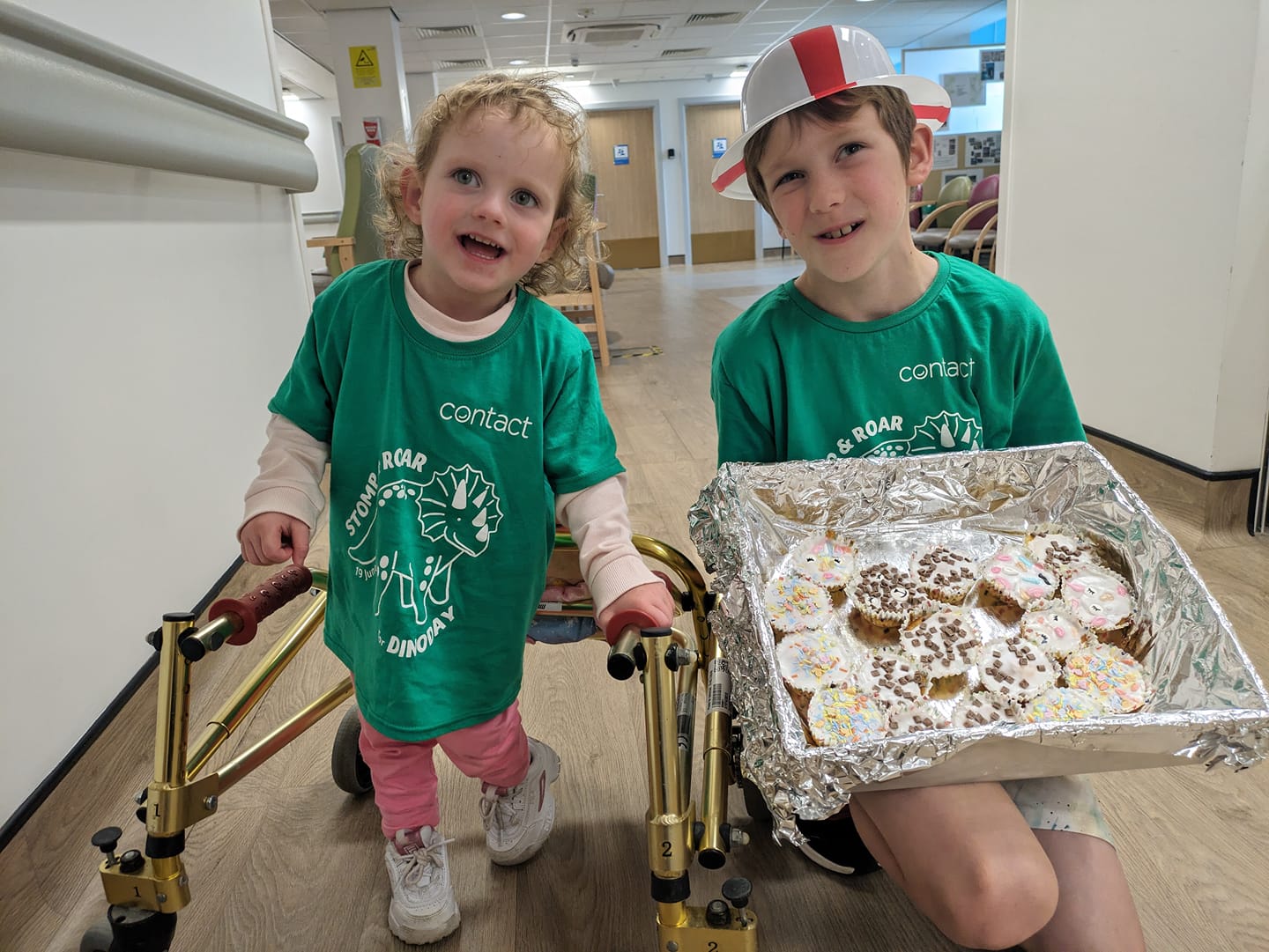 Two young siblings in green t-shirts posing with a box of cupcakes at their local hospital
