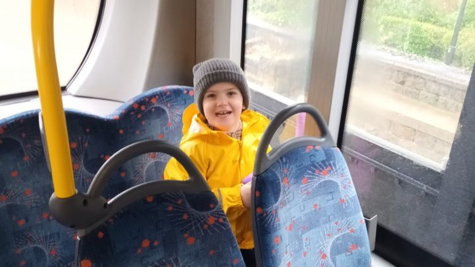 A child sitting on the back row of a public bus smiling at the camera. He is wearing a grey hat and a yellow coat.