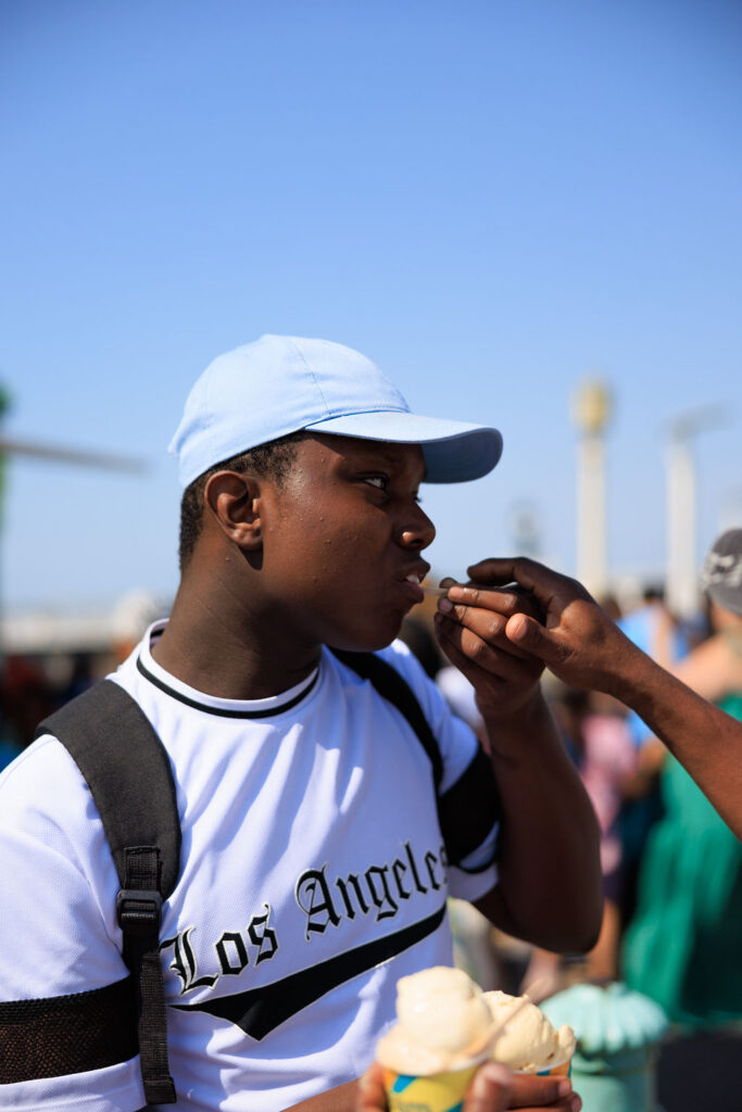 A person wearing a blue cap and "Los Angeles" shirt is being fed ice cream by another hand. They stand outdoors with a group of people in the background, all enjoying a sunny day.