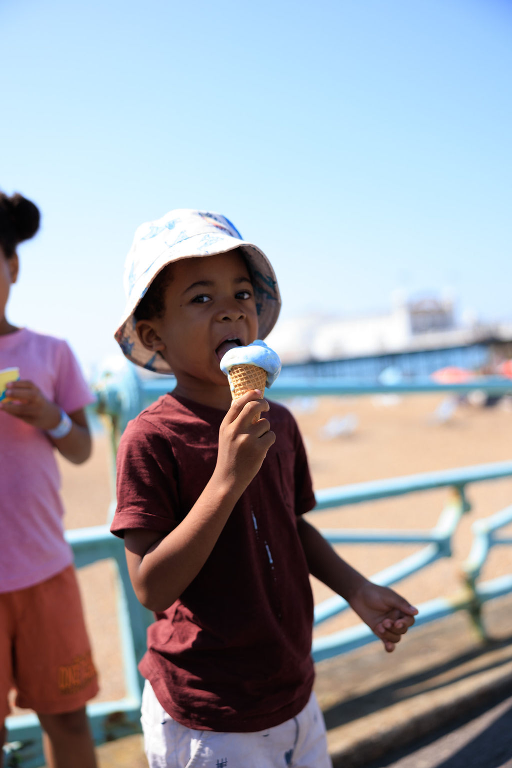 A young boy in a maroon shirt and sun hat is enjoying an ice cream cone on a sunny day. He is standing near a railing with a beach and ocean in the background. Another child is partially visible beside him.