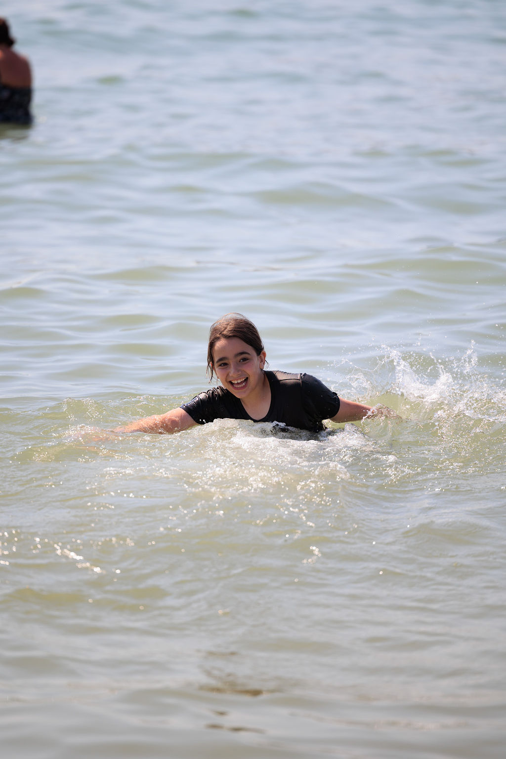 A young girl with long hair, wearing a black shirt, is happily swimming in the ocean. The water is calm, and she is surrounded by gentle waves. Another person is visible in the background. The sky is clear.